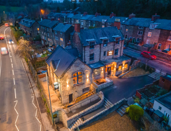 Aerial view of a lit stone pub with surrounding houses at dusk, alongside a curving road with passing cars