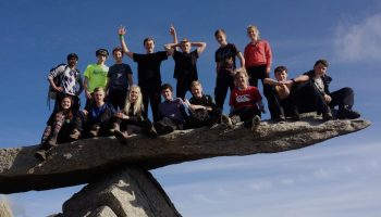 Group of teens sitting and standing on a precarious rock ledge under a clear blue sky during a sunny day outing