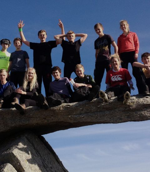 Group of teens sitting and standing on a precarious rock ledge under a clear blue sky during a sunny day outing