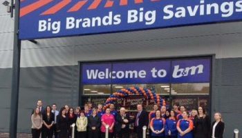 Team of store employees standing in front of B&M retail store entrance beneath a sign saying Big Brands Big Savings