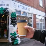 Person holding coffee cup outside Kitchen HQ cafe entrance with balloon arch decoration