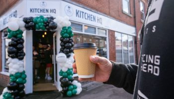 Person holding coffee cup outside Kitchen HQ cafe entrance with balloon arch decoration