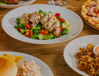Assorted dishes on a wooden table, featuring two pizzas, grilled chicken salad, burger with fries, calamari, and a cocktail