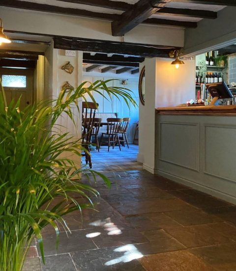 Cosy pub interior with wooden beams, lush green plant, and a classic bar counter leading to a rustic dining area