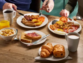 Two people enjoying a full English breakfast with tea, coffee, croissants, toast, and juice at a wooden table