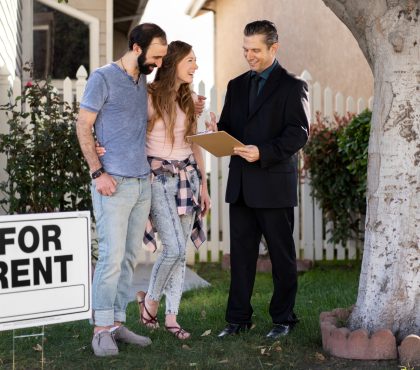 A couple stands in front of a "For Rent" sign, appearing hopeful as they consider their housing options