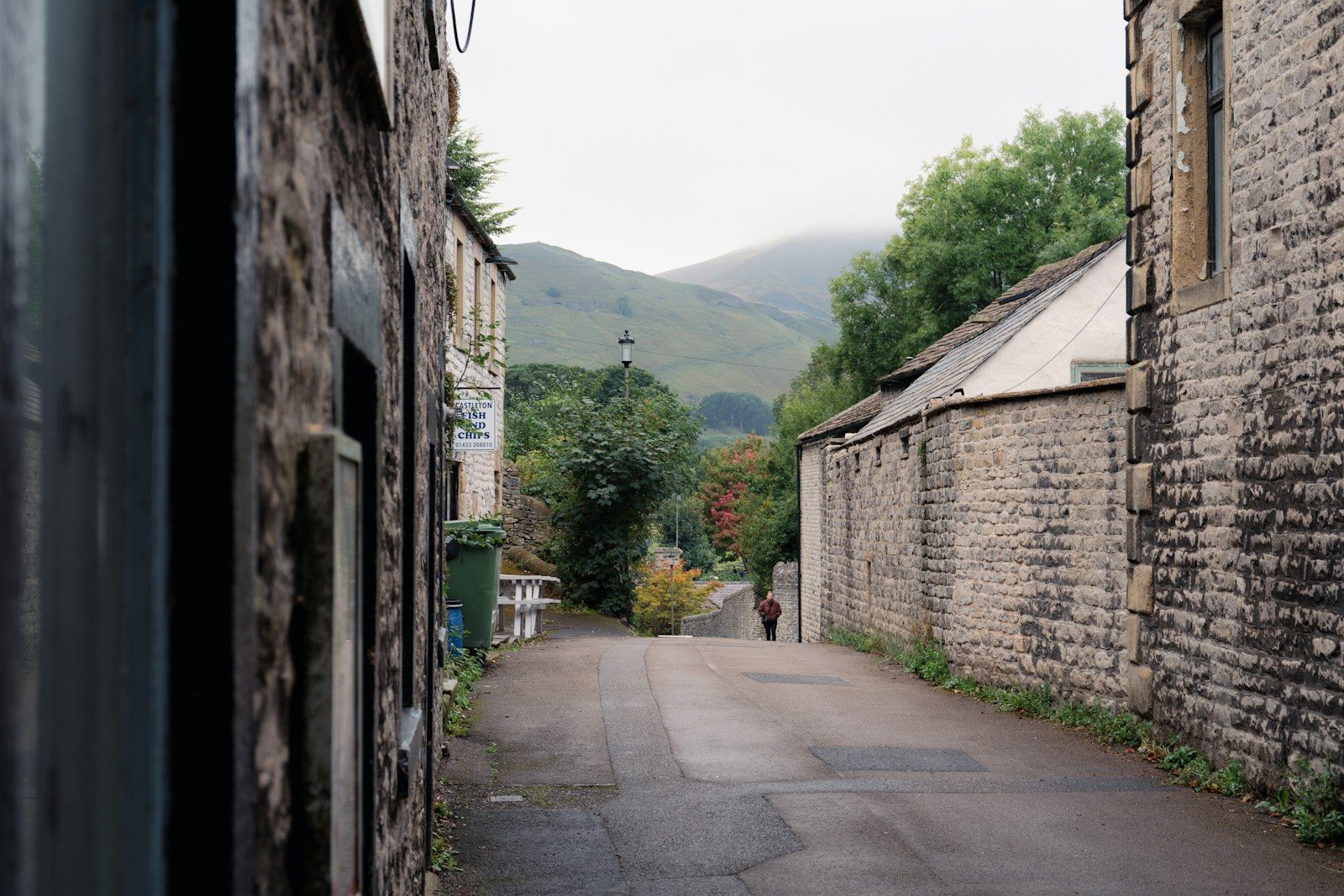 Narrow village lane flanked by stone buildings, leading to rolling green hills under a cloudy sky in rural England
